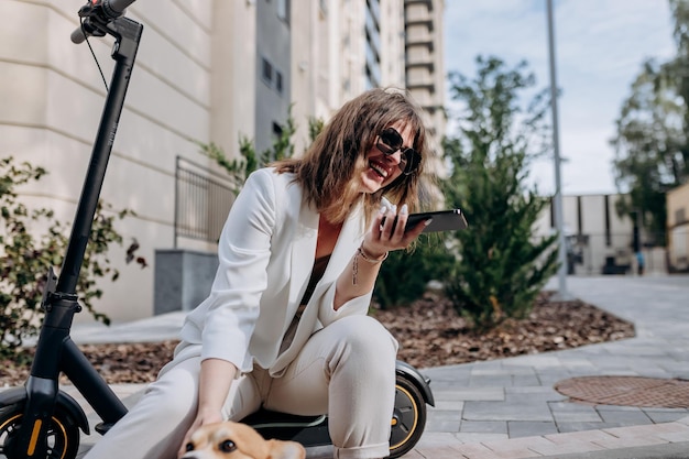 Smiling businesswoman in white suit sitting on electric scooter and records an audio message during walking in city