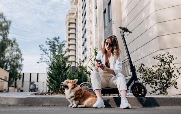 Smiling businesswoman in white suit sitting on electric scooter and using phone during walking with Welsh Corgi Pembroke dog in city