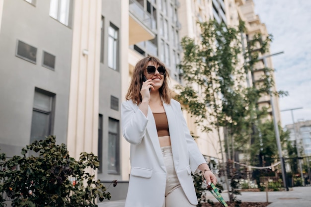 Smiling businesswoman in white suit talking phone during walking in city with modern architecture