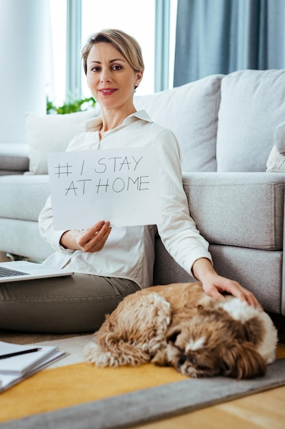 Smiling businesswoman with a dog holding placard with I stay at home hashtag while working at home