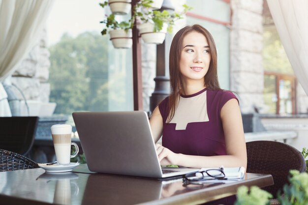 Smiling businesswoman working with laptop sitting at summer terrace cafe. Lifestyle portrait of young business woman. Technology and online communication concept