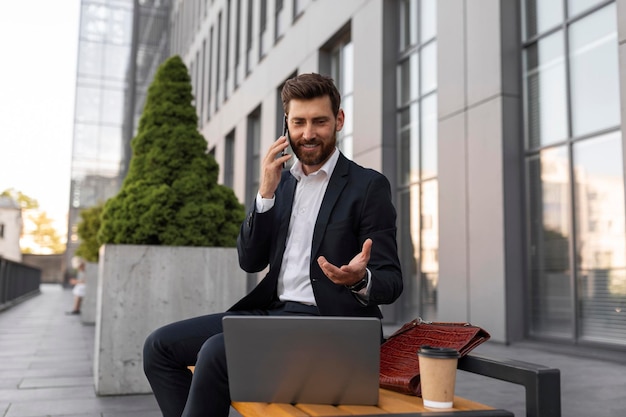 Smiling busy young european man manager with beard in suit with cup of coffee and laptop calls by phone