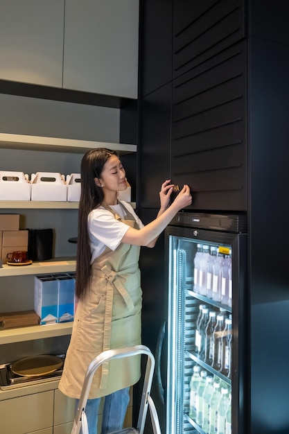 Smiling cafe employee standing on the stepladder while attaching letters to the wall over the refrigerated display cabinet with beverages