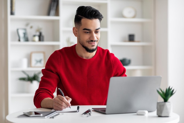 Smiling calm confident attractive millennial arab guy with beard in red clothes makes notes works on