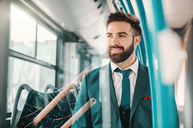 Smiling Caucasian bearded businessman in turquoise suit sitting in public bus and looking trough window. Good energy is contagious.