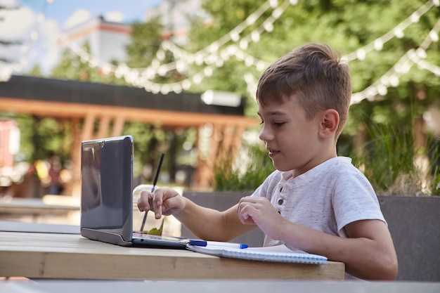 Smiling caucasian boy sitting at table at veranda of cafe studying with laptop