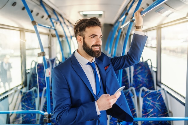 Smiling caucasian businessman in formal wear standing in public bus, holding bar and using smart phone.