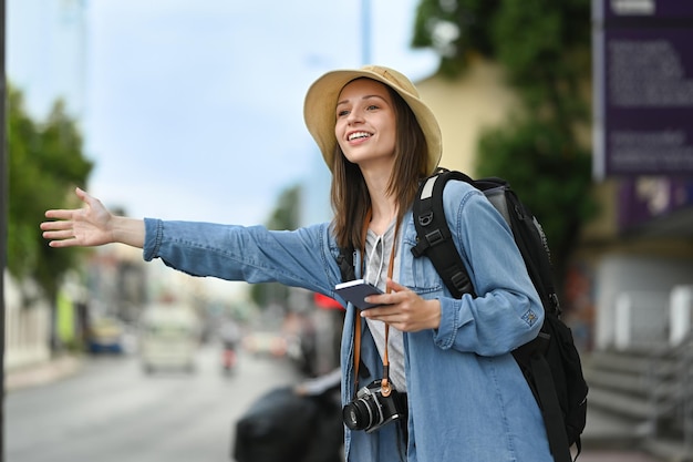 Smiling caucasian female traveler raising hand calling taxi in busy city street Tourism and transportation concept