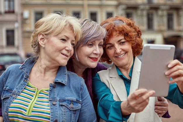 Smiling caucasian middle-aged females are taking selfie by digital device on open air.