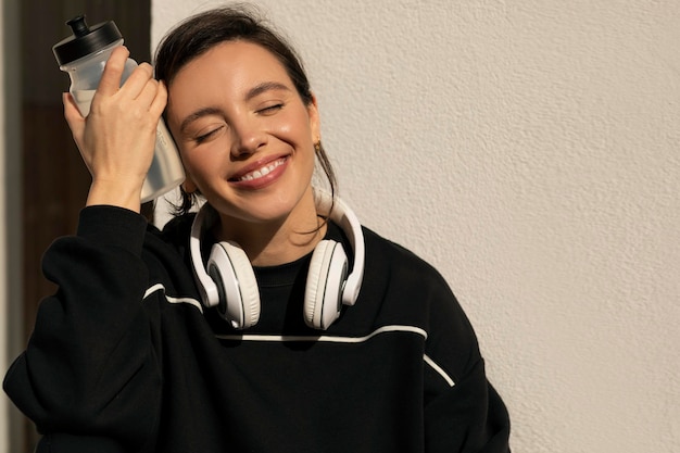 Smiling caucasian millennial lady in sportswear and headphones relaxing with bottle of water on street