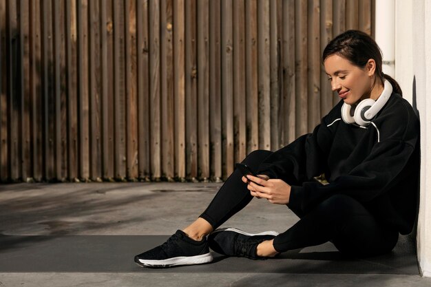 Smiling caucasian millennial lady in sportswear and headphones resting sitting on floor typing on smartphone