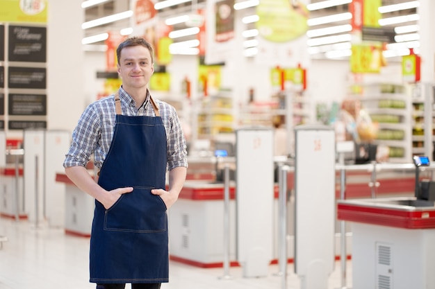 Smiling caucasian seller stands holding hands in pockets with superstore