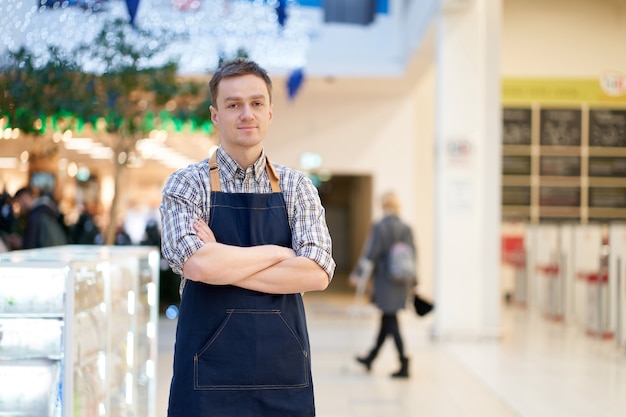 Smiling caucasian seller strands with crossed hands, superstore. Copy space.