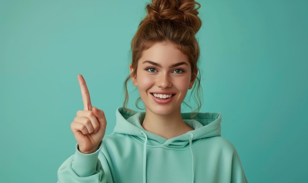 Photo smiling caucasian teenager girl in a mint sweatshirt smiling with healthy teeth and pointing index finger up at copy space expressing wow emotion standing isolated on paper mint background