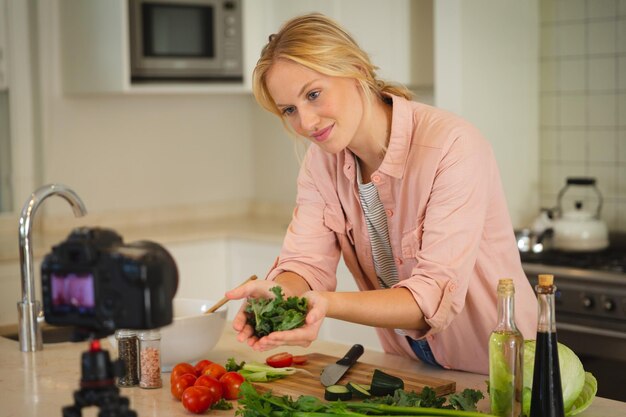 Smiling caucasian woman in kitchen holding vegetables and looking at camera, making cooking vlog. technology and communication, cookery vlogger at home.