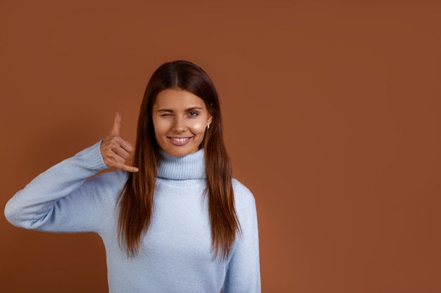 Smiling caucasian woman wearing blue sweater makes telephone gesture, asks to call her, blinks eyes