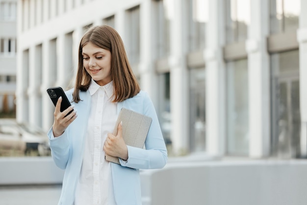 Smiling caucasian woman wearing formal suit using mobile phone typing text messages walking holding