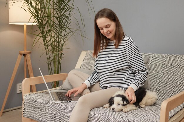 Smiling Caucasian woman wearing striped shirt sitting on sofa with her puppy dog using laptop comput