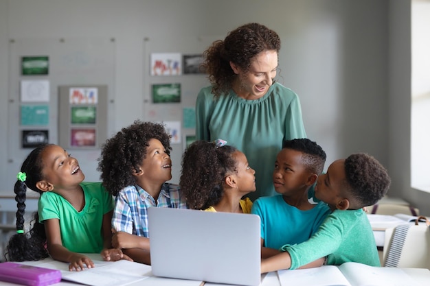 Smiling caucasian young female teacher teaching laptop to african american elementary students