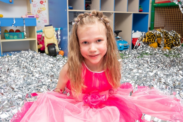 Smiling cheerful girl in a pink dress in a huge pile of silver foil confetti in the nursery A girl in sparkling sequins