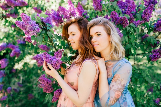 Smiling cheerful twin sisters in stylish dresses posing in summer park.