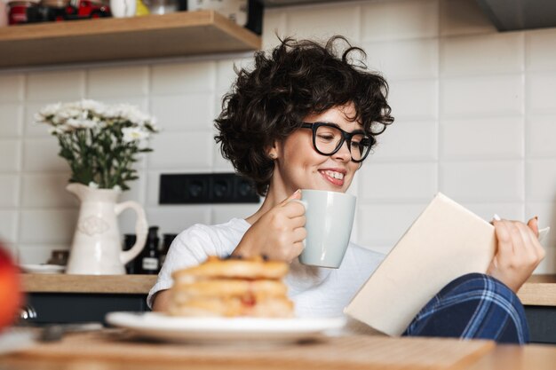 Smiling cheerful woman having tasty breakfast while sitting at the kitchen at home, reading a book