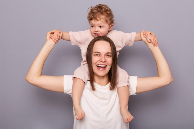 Smiling cheerful woman with brown hair holding her baby daughter on shoulders enjoying to play together posing isolated over gray background Childhood motherhood