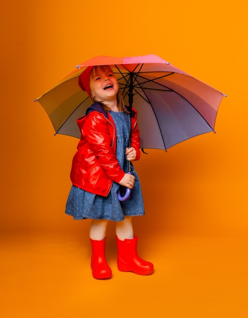 A smiling child girl in a red jacket and boots, holding an umbrella, stands on a yellow background. little girl in bright clothes holds a rainbow umbrella.