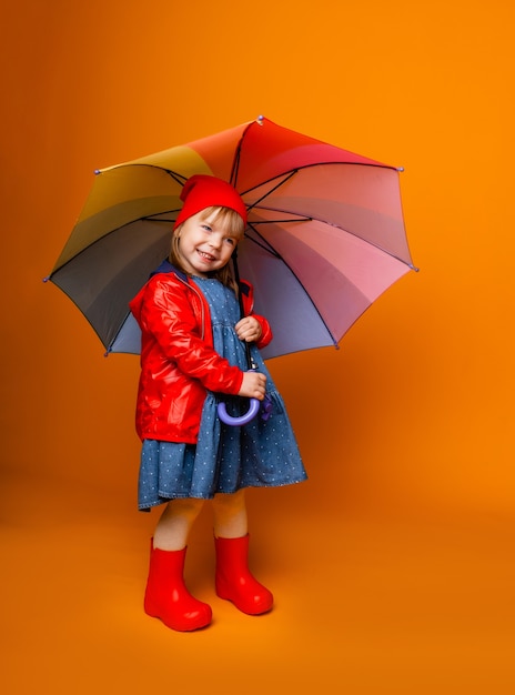 A smiling child girl in a red jacket and boots, holding an umbrella, stands on a yellow background. little girl in bright clothes holds a rainbow umbrella.