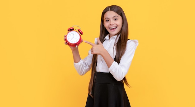 Smiling child in school uniform point finger on alarm clock showing time on yellow background back to school