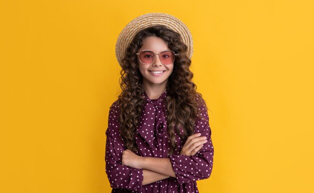 Smiling child in straw hat and sunglasses with long brunette curly hair on yellow background