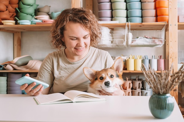 Smiling concentrated businesswoman holding smartphone with cute dog corgi in workshop Silicone baby dishes shop