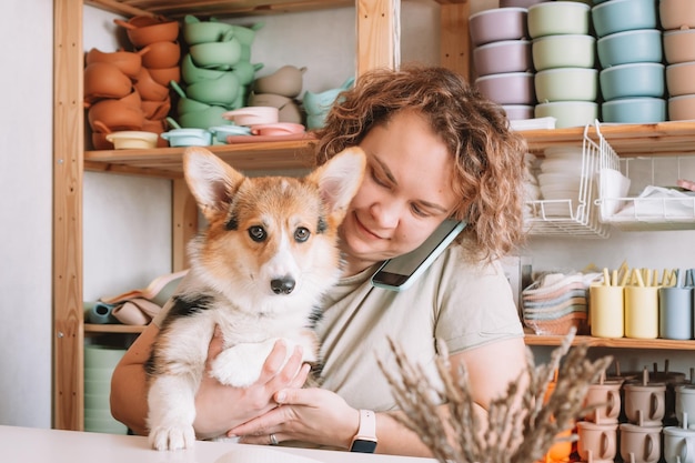 Smiling concentrated businesswoman talking smartphone holding cute dog corgi in workshop Silicone baby dishes shop