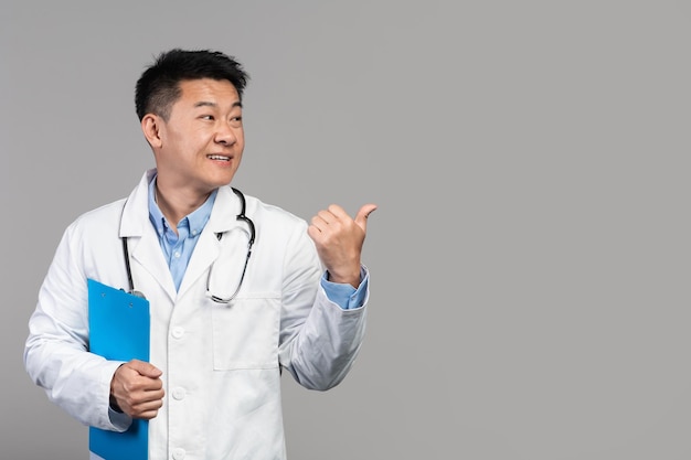 Smiling confident adult asian doctor in white coat with stethoscope and tablet pointing finger at empty space