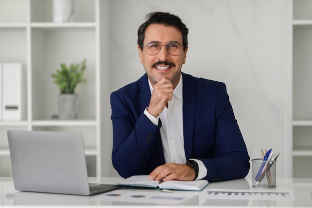 Smiling confident caucasian adult businessman in suit at table with computer laptop