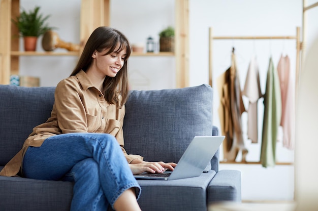 Smiling confident lady entrepreneur in casual outfit sitting on sofa and typing on laptop in own clo