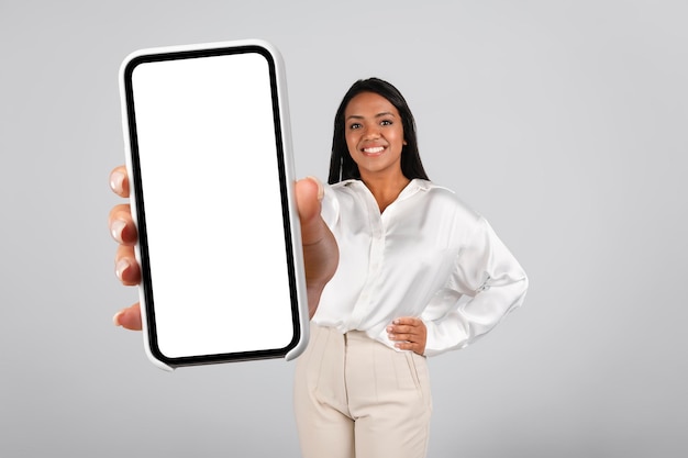 Smiling confident millennial black female in white blouse show phone with empty screen recommend new app