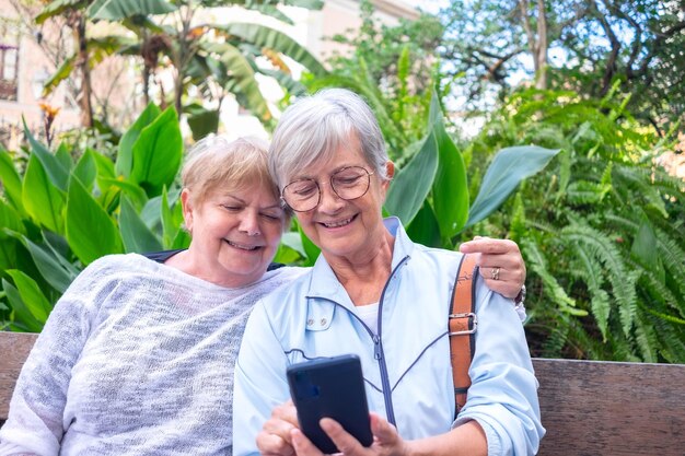 Smiling couple of elderly caucasian women sitting relaxed in the park while looking at the same cellphone Senior females hugging sharing tech and news