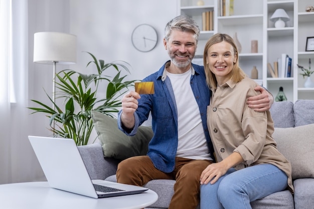 Smiling couple holding a credit card while shopping online with a laptop in their cozy living room