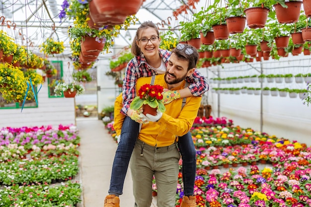 Smiling couple in love having piggyback ride in greenhouse. All around them are colorful flowers. Small business owners.