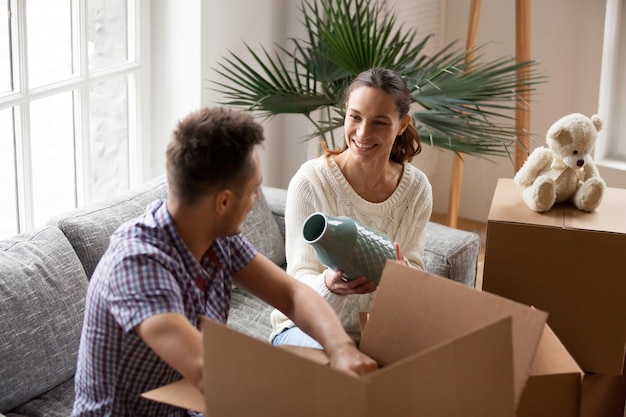 Smiling couple packing cardboard boxes together sitting on sofa in living room preparing to relocate young happy woman holding vase helping man to unpack belongings moving in new home concept