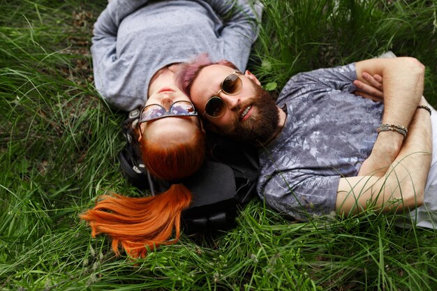 Photo smiling couple, seated down on a green grass, in park, seated down in sunglasses, looking at camera. horizontal view.