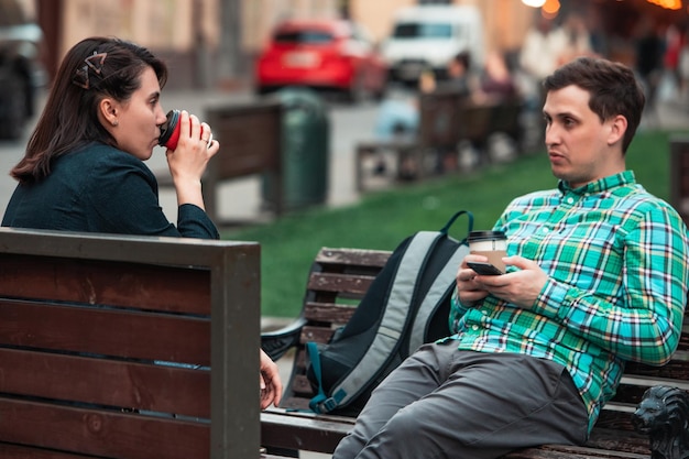 Smiling couple sitting on bench talking to each other drinking coffee friends urban life