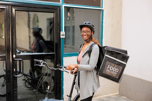 Smiling courier on bike looking at camera, waiting for customer near office building entrance outdoors. Restaurant takeaway meal delivery service, african american woman standing in front of door