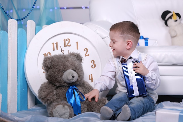 Smiling cute boy holding his present during christmas time. big white clock and teddy bear on wall