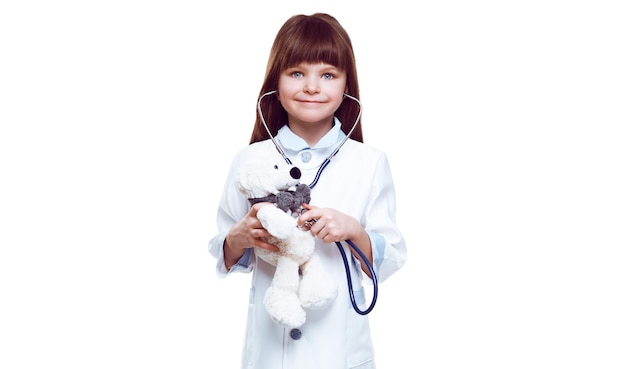 Smiling cute caucasian girl playing doctor and listening teddy bear with stethoscope isolated on white background