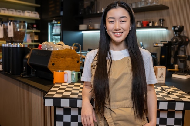 Smiling cute longhaired young Asian female employee leaning on the counter in the coffee shop