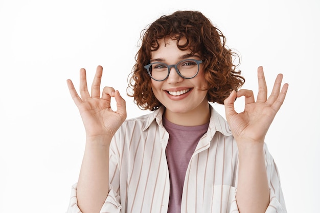 Smiling cute queer girl in glasses showing okay ok gesture, looking assured and confident in her choice, guarantee and recommend advertisement, like and approve something good.