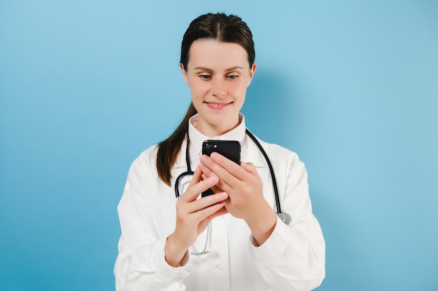 Smiling cute woman doctor in white coat standing isolated on blue background hold smartphone, check messages from patient, use mobile app for medical workers easy comfort usage of modern tech concept