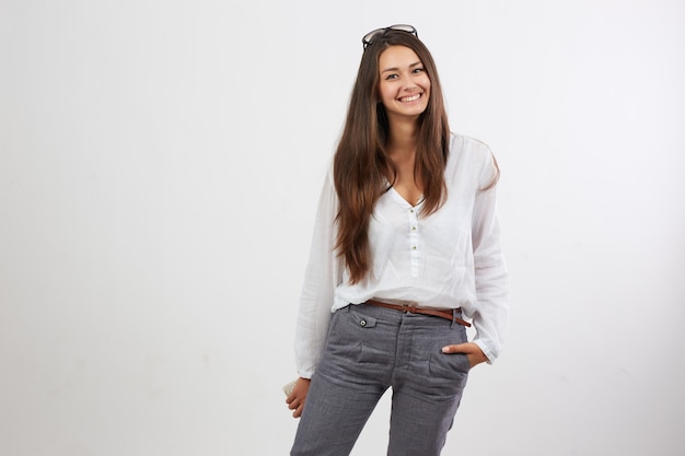 Smiling dark-haired dressed in white blouse and gray trousers stands on the white background .